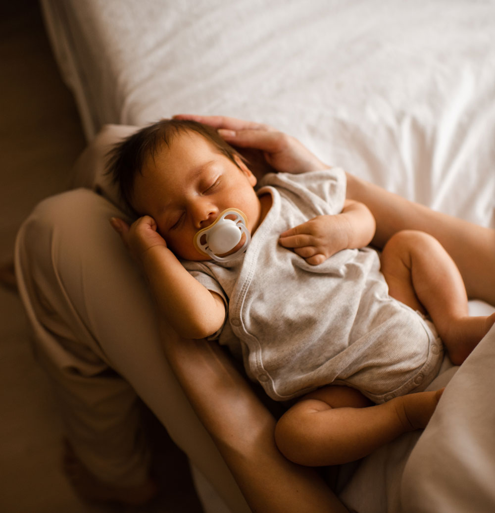 Sleeping baby with pacifier resting in parent's arms on bed.