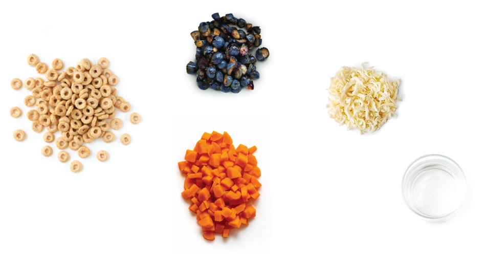 Top-down view of food ingredients including dry cereal rings, blueberries, shredded cheese, diced carrots, and a glass of water on a white background