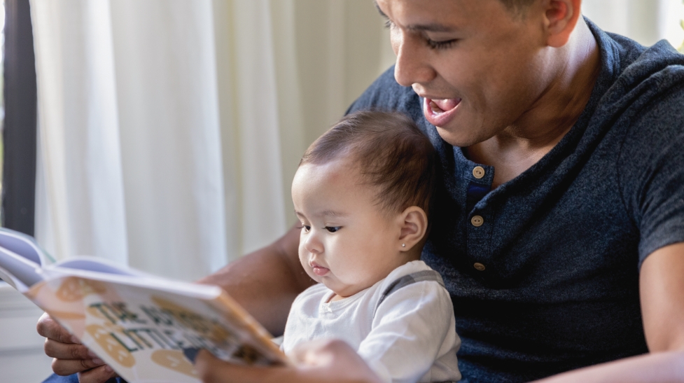 A father reading a book to his baby, possibly holding the baby in his lap or next to him.