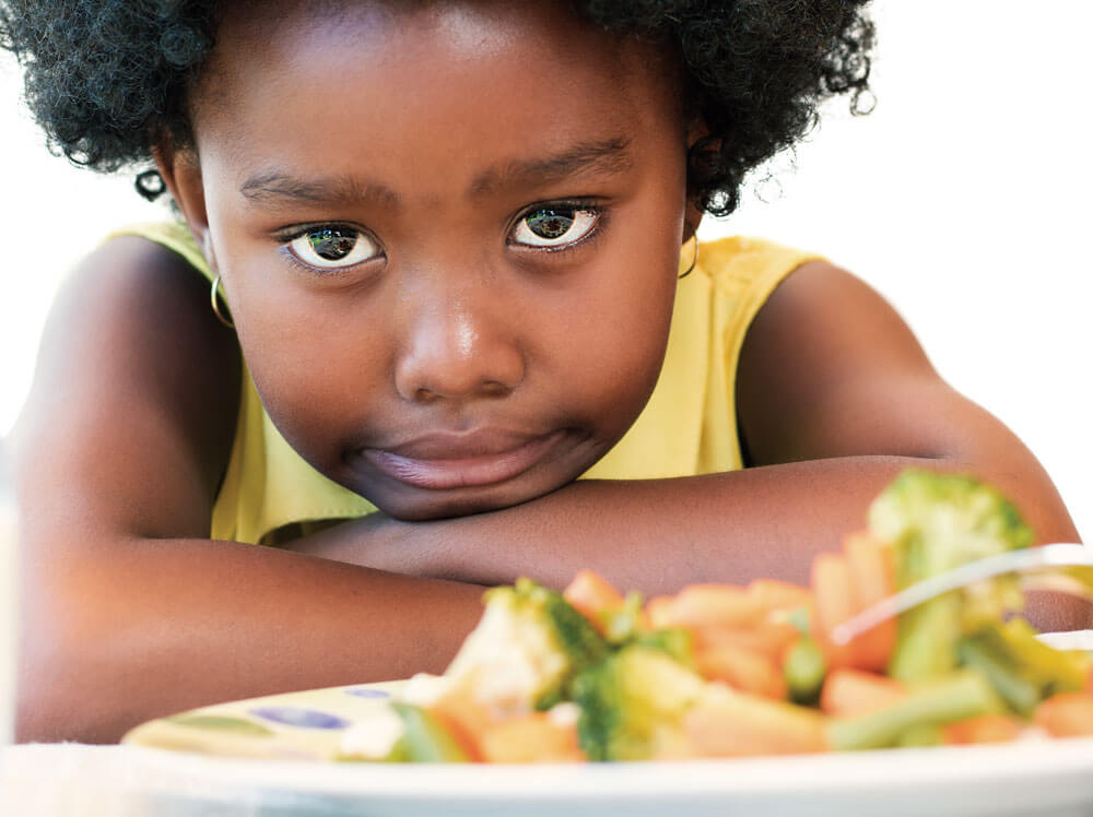 "A young girl with a grumpy expression sitting at a table with food in front of her.