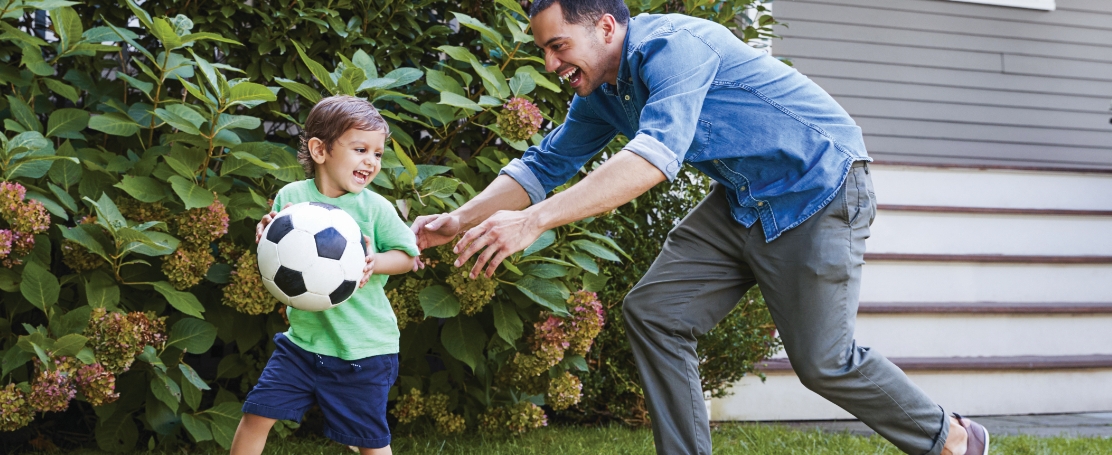 A father and son playing soccer together outdoors, with the father possibly coaching or just having fun with his son. The child is likely kicking the ball or trying to make a move, while the father is engaged and encouraging.