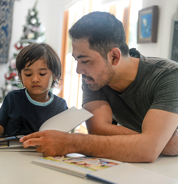 A father reading a book to his young son, with both sitting together in a cozy, engaging moment. The father is likely holding the book, while the son is looking at the pages, showing interest and attentiveness.